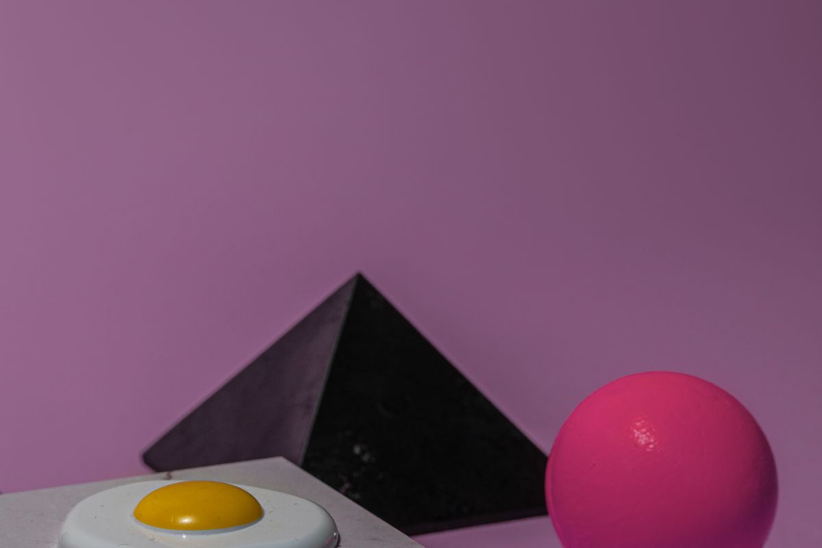 A black pyramid, a pink sphere and an egg yolk arranged on a purple background.