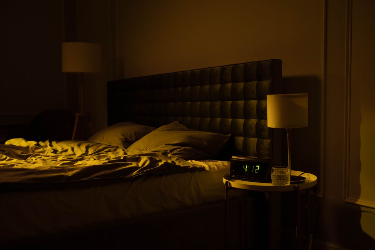 A dimly lit bedroom at night, an unmade bed, and a glowing digital clock on the nightstand.